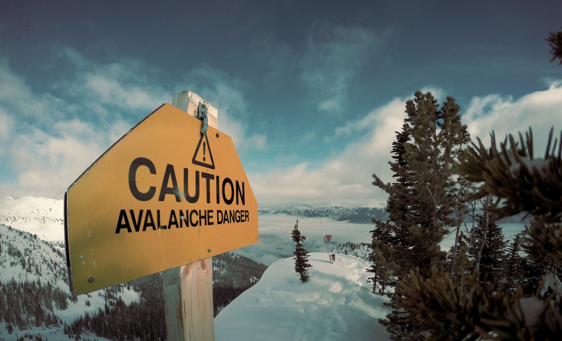 Photo of a snowy mountain landscape. Prominent in the foreground is a yellow warning sign which says "CAUTION, AVALANCHE DANGER"